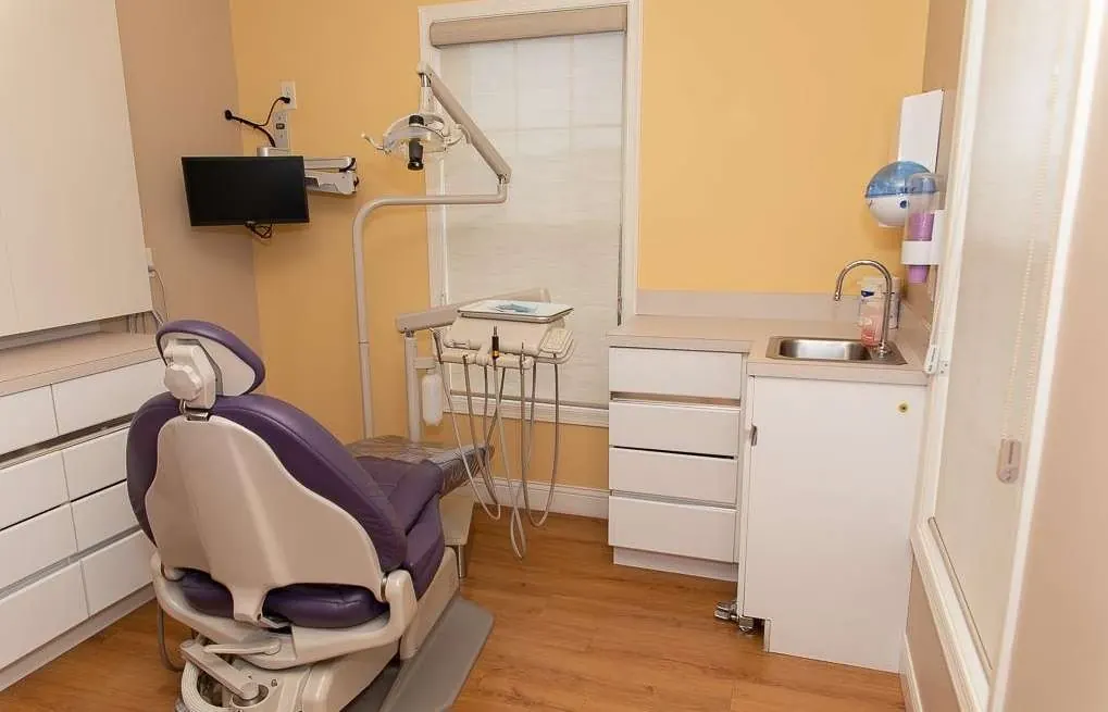 A treatment room with a purple dental chair, cabinets and a wall-mounted screen.